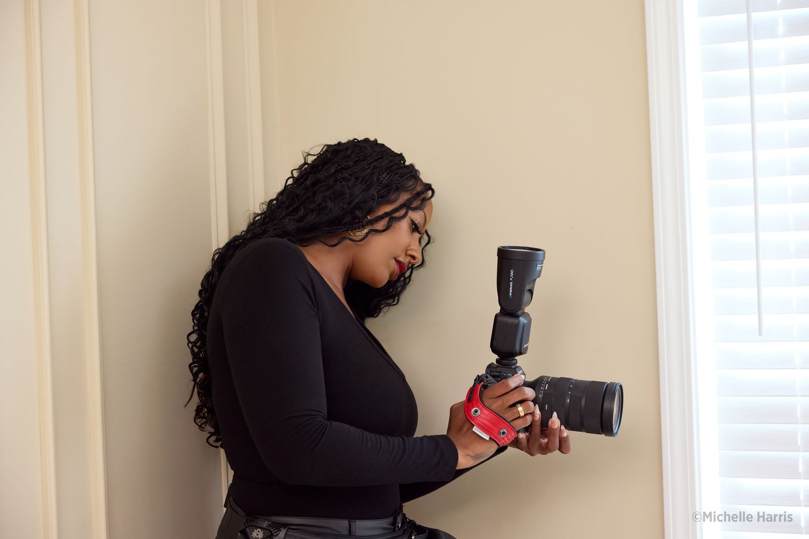 A woman with long dark braided hair wearing a black long-sleeved shirt holds a camera with a mounted flash while looking down at the screen.