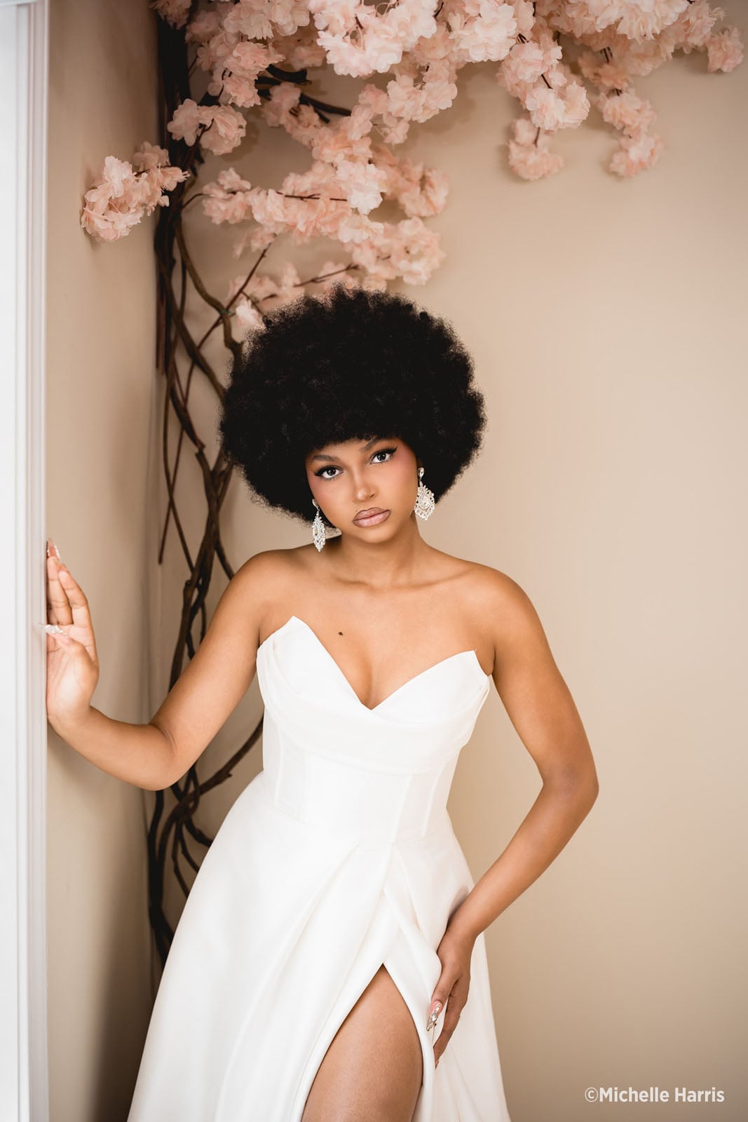 A woman with a large afro hairstyle wears a strapless white gown and crystal earrings while posing next to a flowering cherry blossom branch.