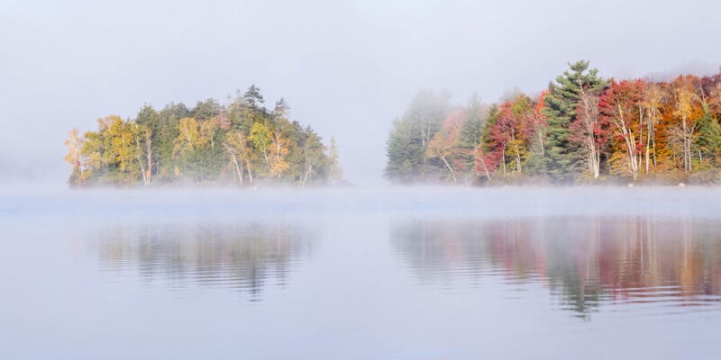 A calm lake reflects a misty shoreline featuring trees with autumn foliage in shades of yellow, orange, and red.