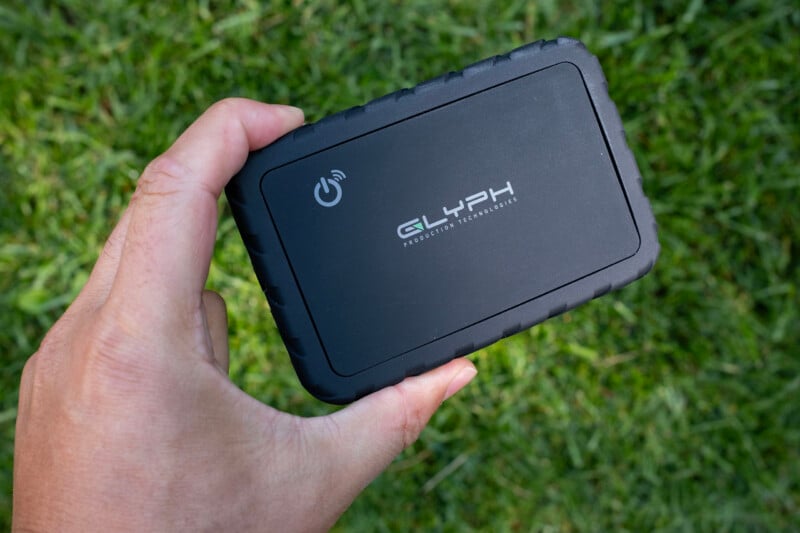 A hand holds a black, rectangular Glyph Production Technologies external hard drive against a background of green grass.