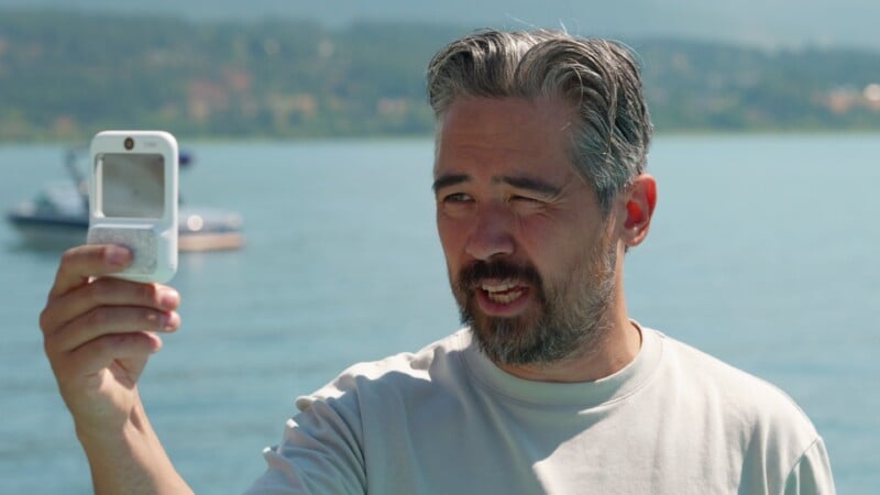 A middle-aged man with graying hair and a beard squints while holding a small, white rectangular device in front of his face outdoors by a lake.