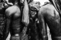 A young child with a dark, painted face and head covering stands between two adults in the rain, all with wet, glistening skin.