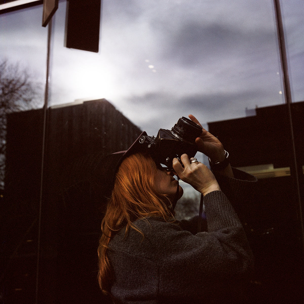 A person with long red hair wearing a dark sweater and hat holds a medium-format camera up to their eye to take a picture.