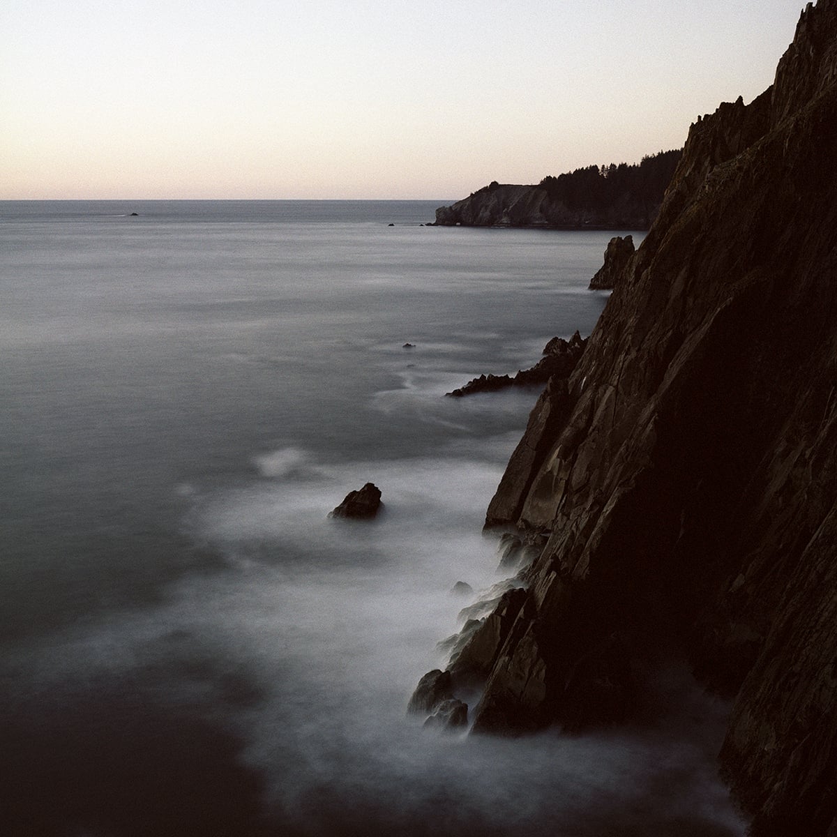A long-exposure view of a rocky coastline at dusk, with soft, blurred waves washing against dark cliffs.