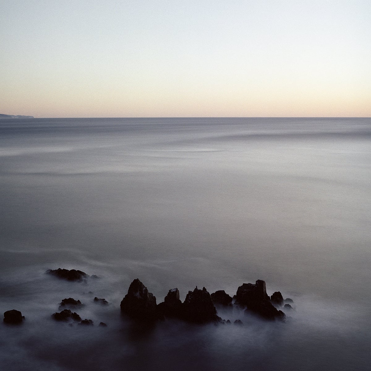 A long-exposure shot of dark, jagged rocks emerging from a smooth, misty ocean under a pale, twilight sky.
