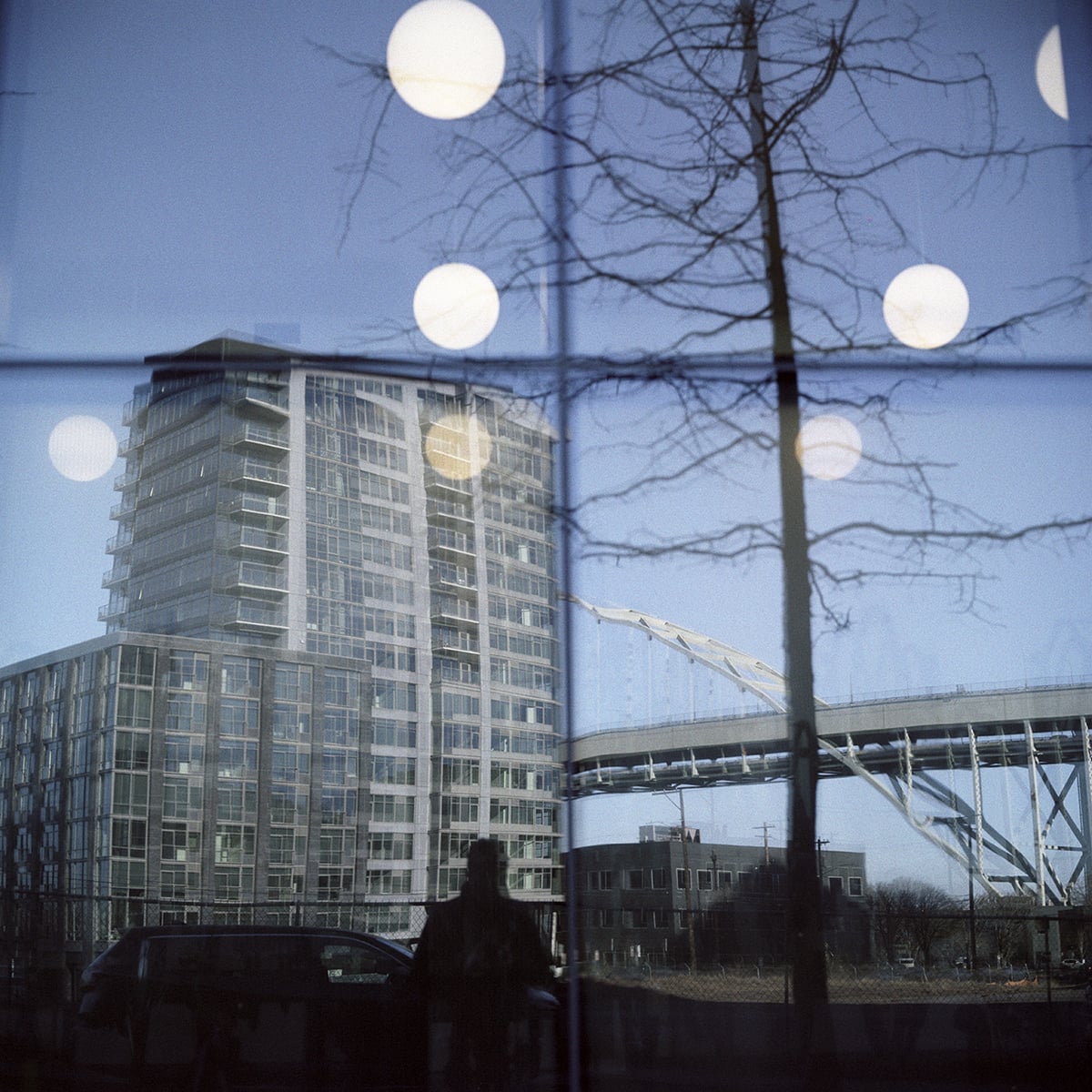 A reflection in a window shows a tall apartment building, a large bridge, and a bare tree, overlaid with glowing circular light fixtures.