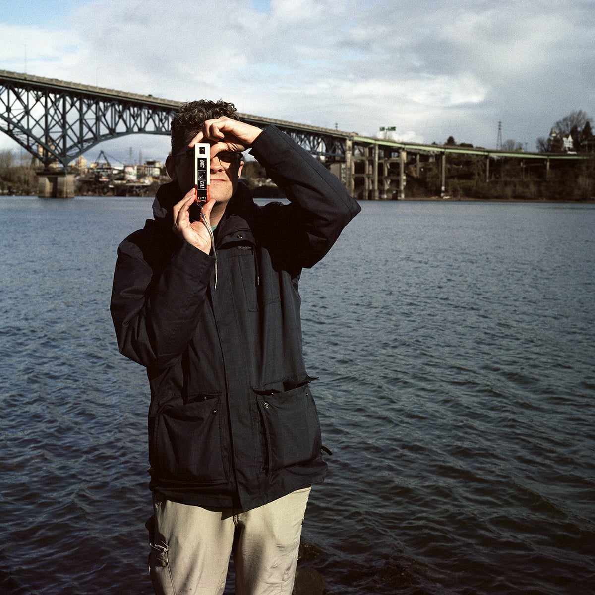 A person in a dark jacket and light pants holds a small, rectangular camera up to their eye in front of a large bridge over a river.