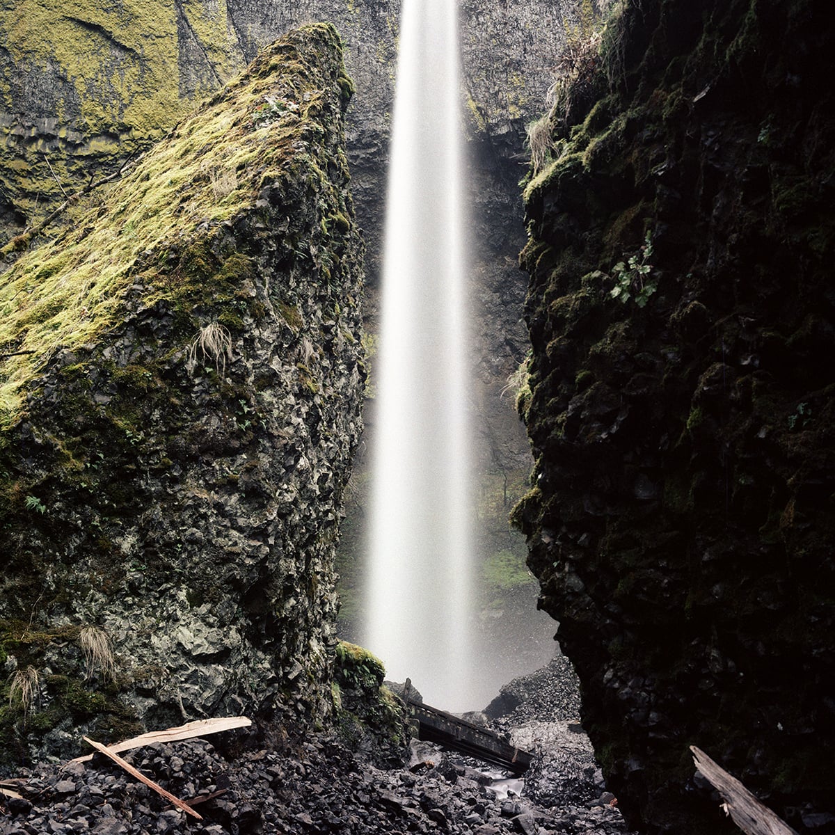 A tall, narrow waterfall cascades between two large, moss-covered rock formations over a dark, rocky ground.