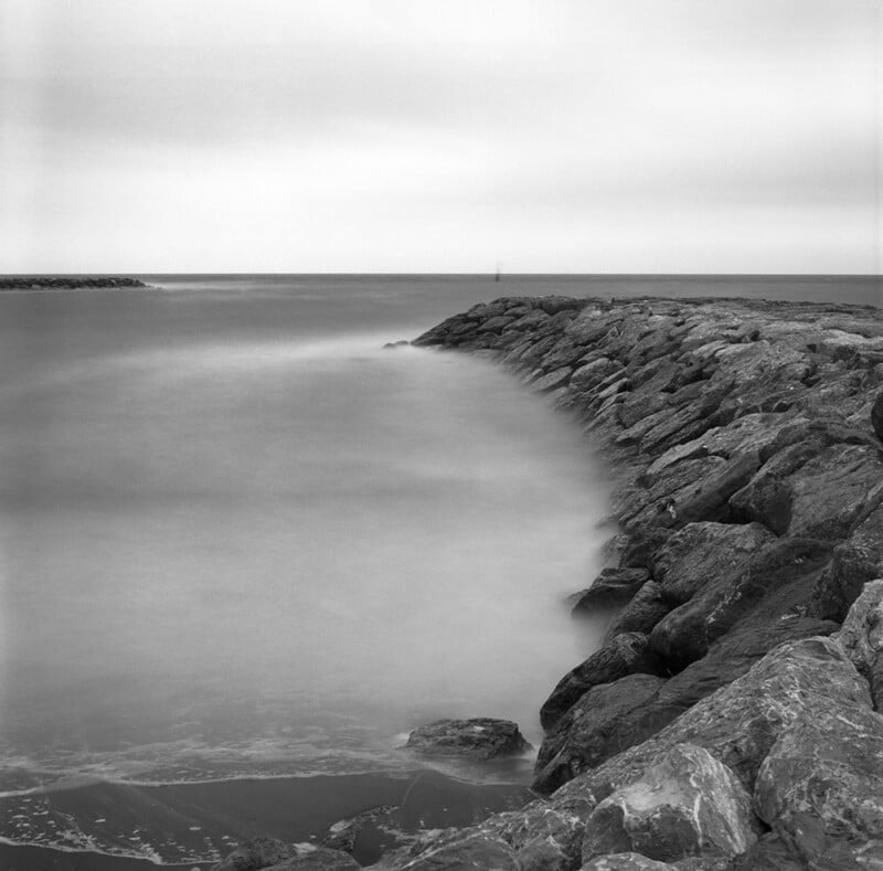 A long exposure black and white shot of a rocky jetty extending into a calm, misty sea under an overcast sky.
