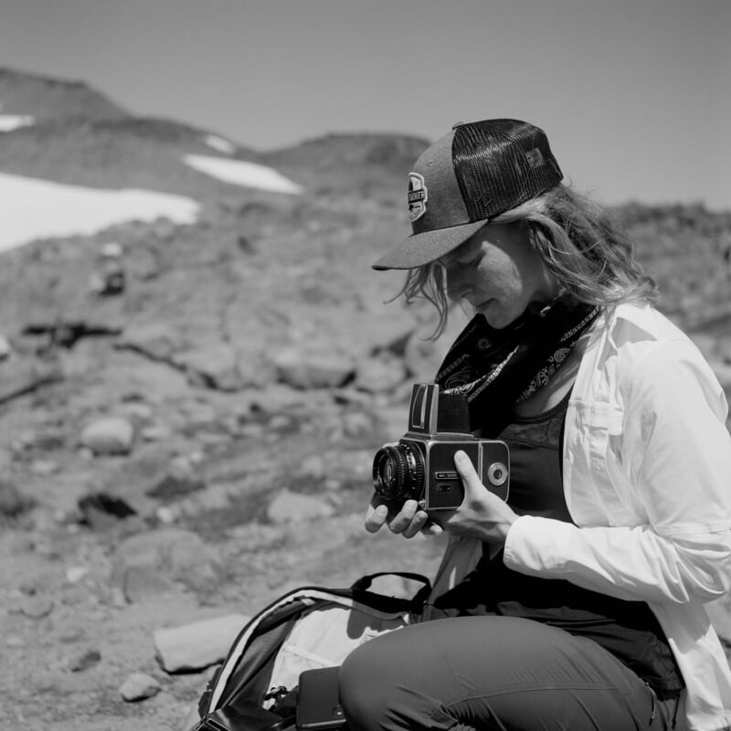 A woman wearing a baseball cap and a bandana around her neck holds a medium format camera while sitting outdoors in a rocky, mountainous landscape.