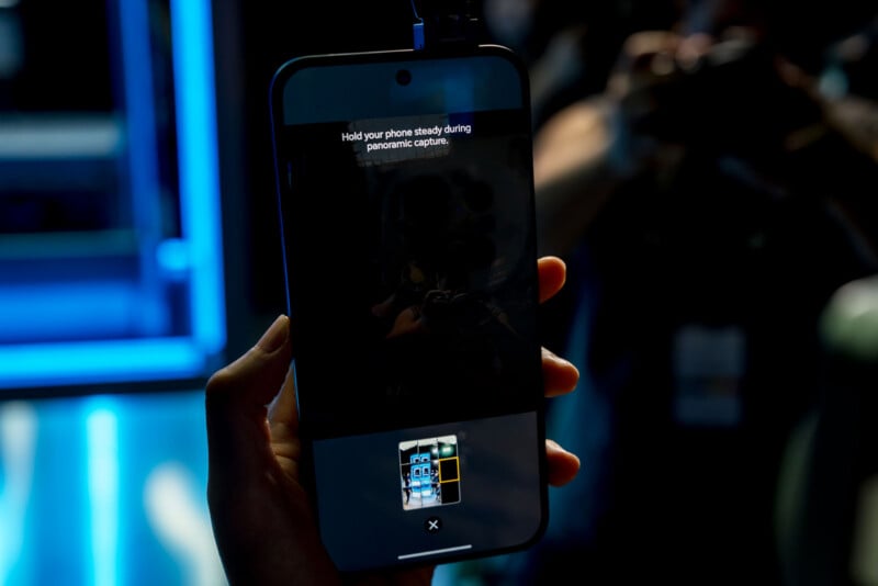 A hand holds a smartphone displaying a panoramic capture interface with the instruction to hold the phone steady.