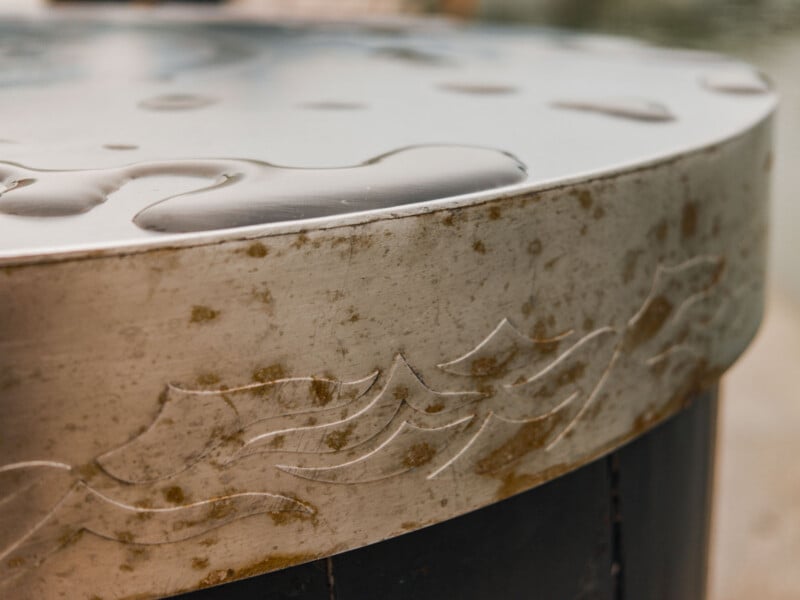 A close-up view of a circular metal table edge featuring an engraved wave pattern and water droplets on the surface.