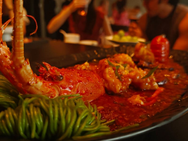 A large, cooked lobster served in a spicy red sauce with a side of green noodles on a dark platter.