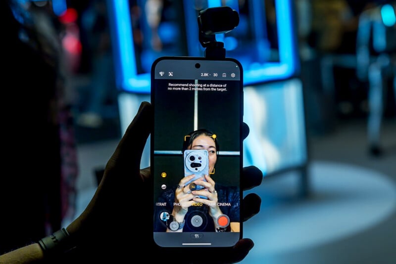 A hand holds a smartphone displaying a camera interface with a prompt to shoot within two meters of the target, while a small gimbal camera is attached to the top of the phone.