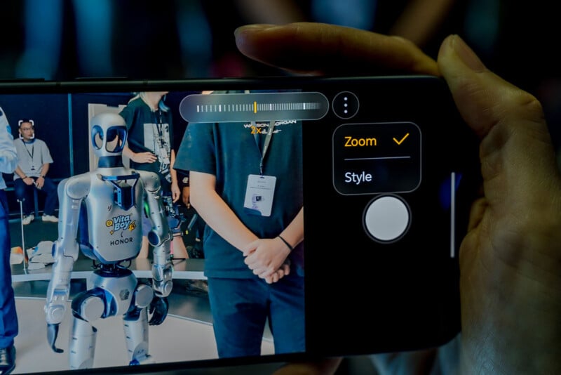 A hand holds a smartphone displaying a camera interface with a 2x zoom setting, showing a silver robot and a person in the background.