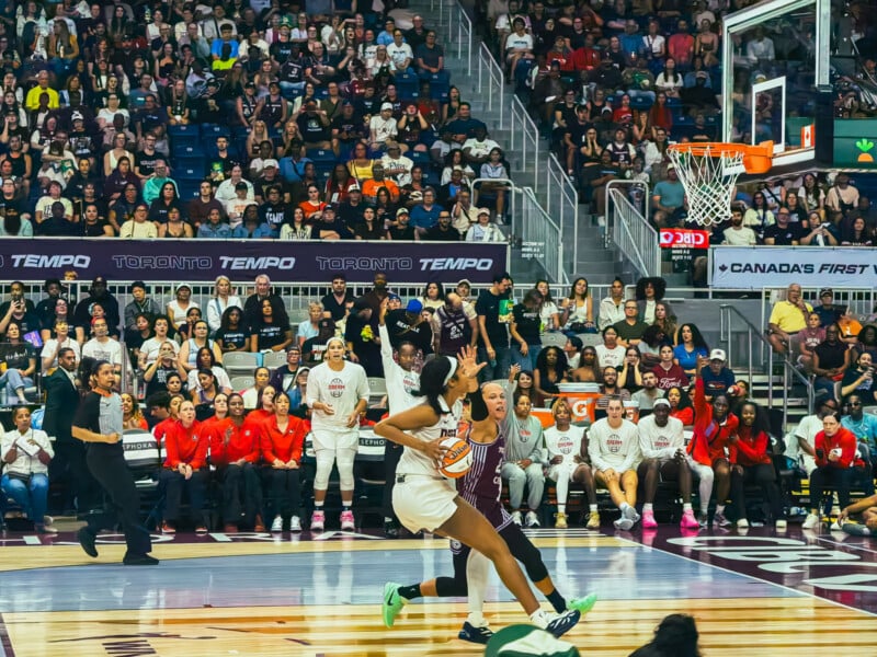 A basketball player in a white uniform dribbles the ball past a defender in a maroon uniform during a game in a crowded arena.