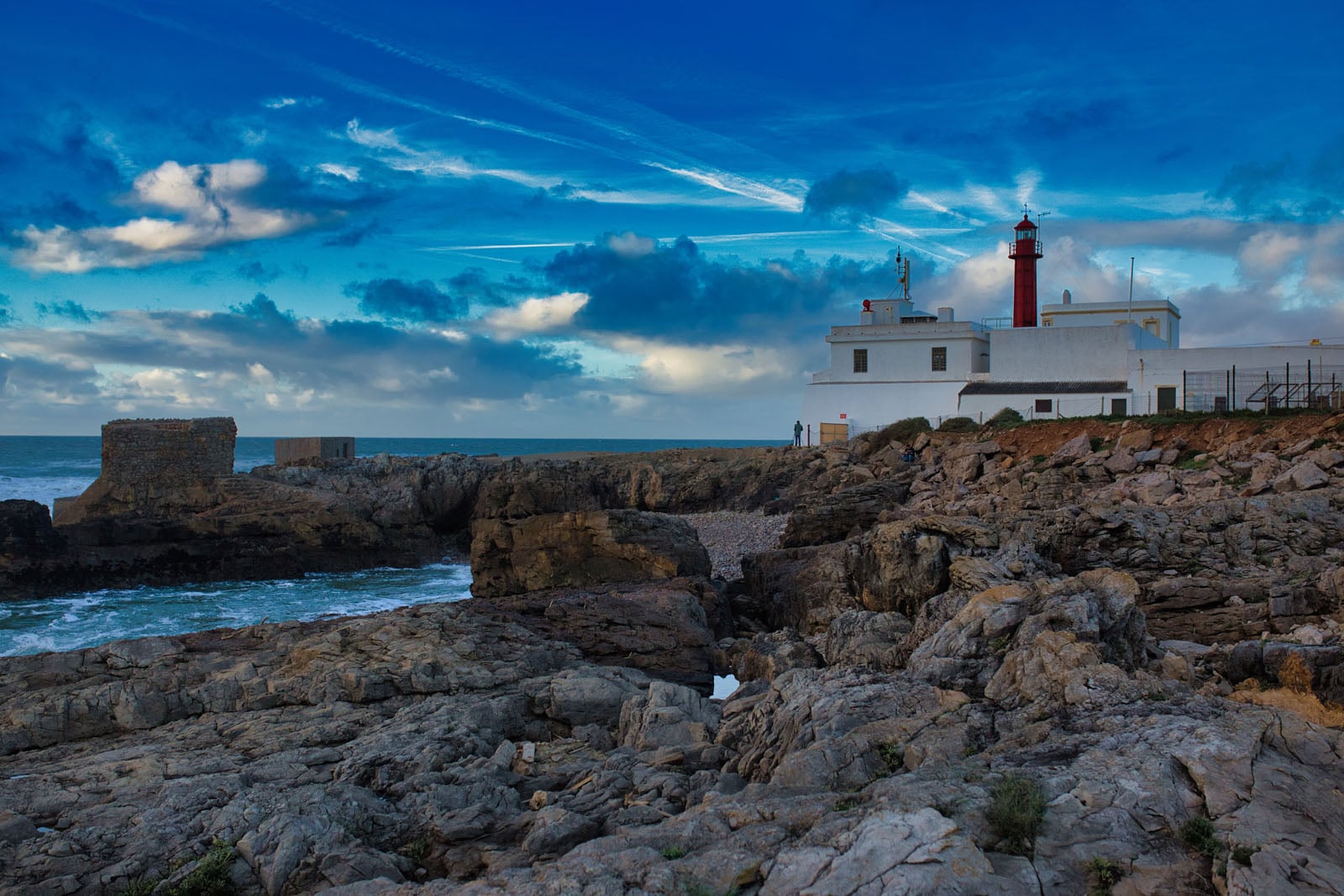 A white lighthouse with a red lantern room stands on a rocky coastline under a blue sky with scattered clouds.