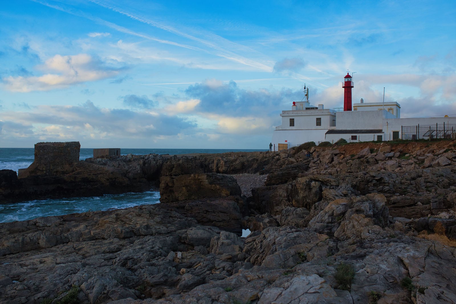 A white lighthouse with a red lantern room stands on a rocky coastline overlooking the ocean under a blue, cloudy sky.