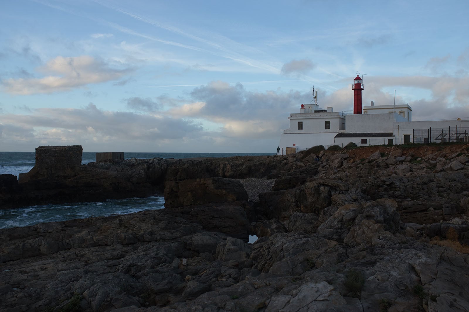 A white lighthouse with a red lantern room stands on a rocky coastline next to the ocean under a cloudy sky.