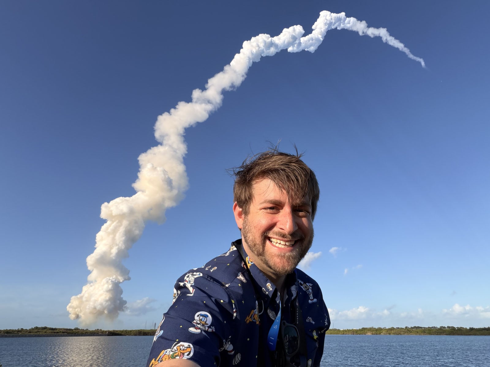 A smiling man wearing a patterned shirt poses for a selfie in front of a rocket launch plume rising into a clear blue sky.