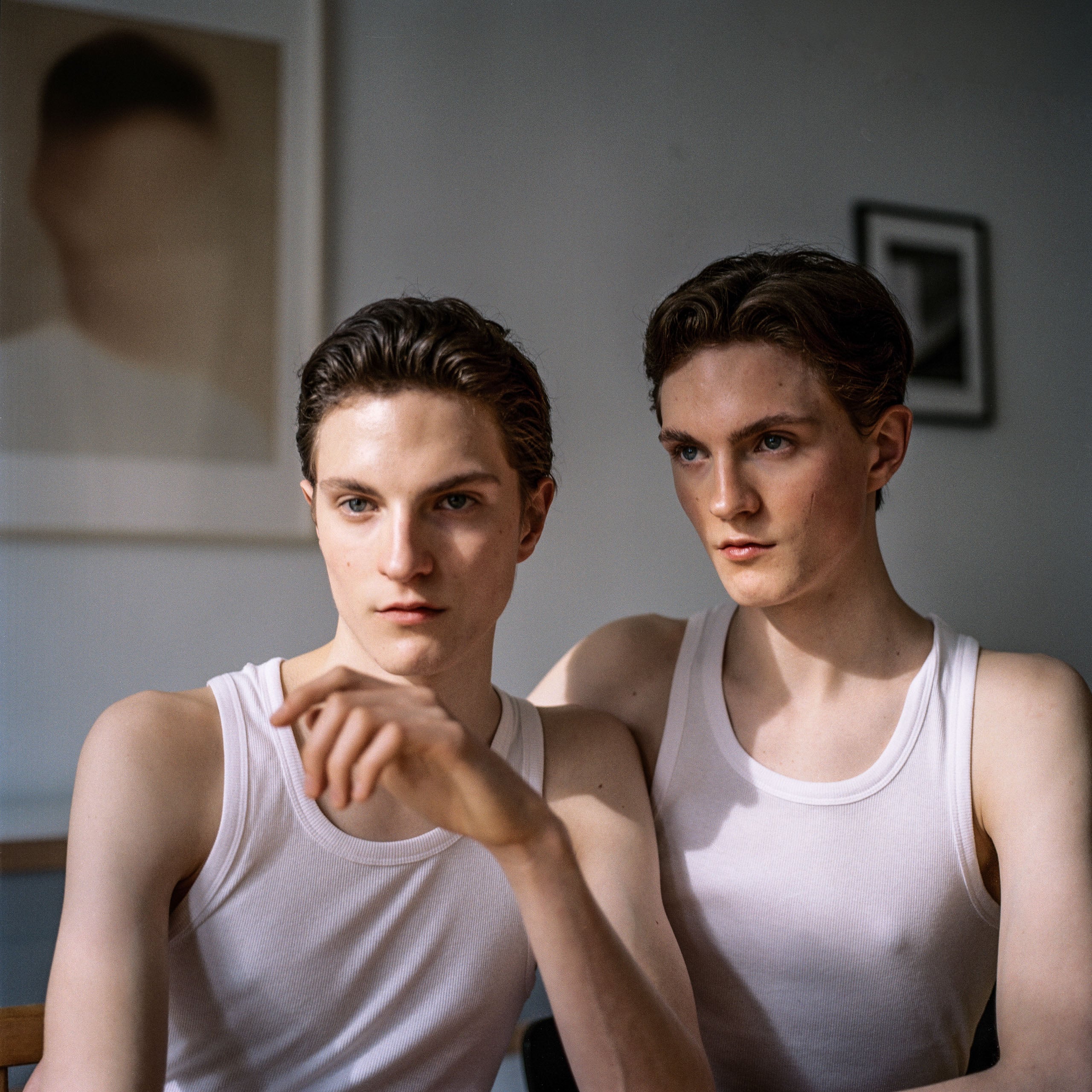 Two young men with similar features and dark hair wear white tank tops, sitting side-by-side in a room with framed art on the wall.