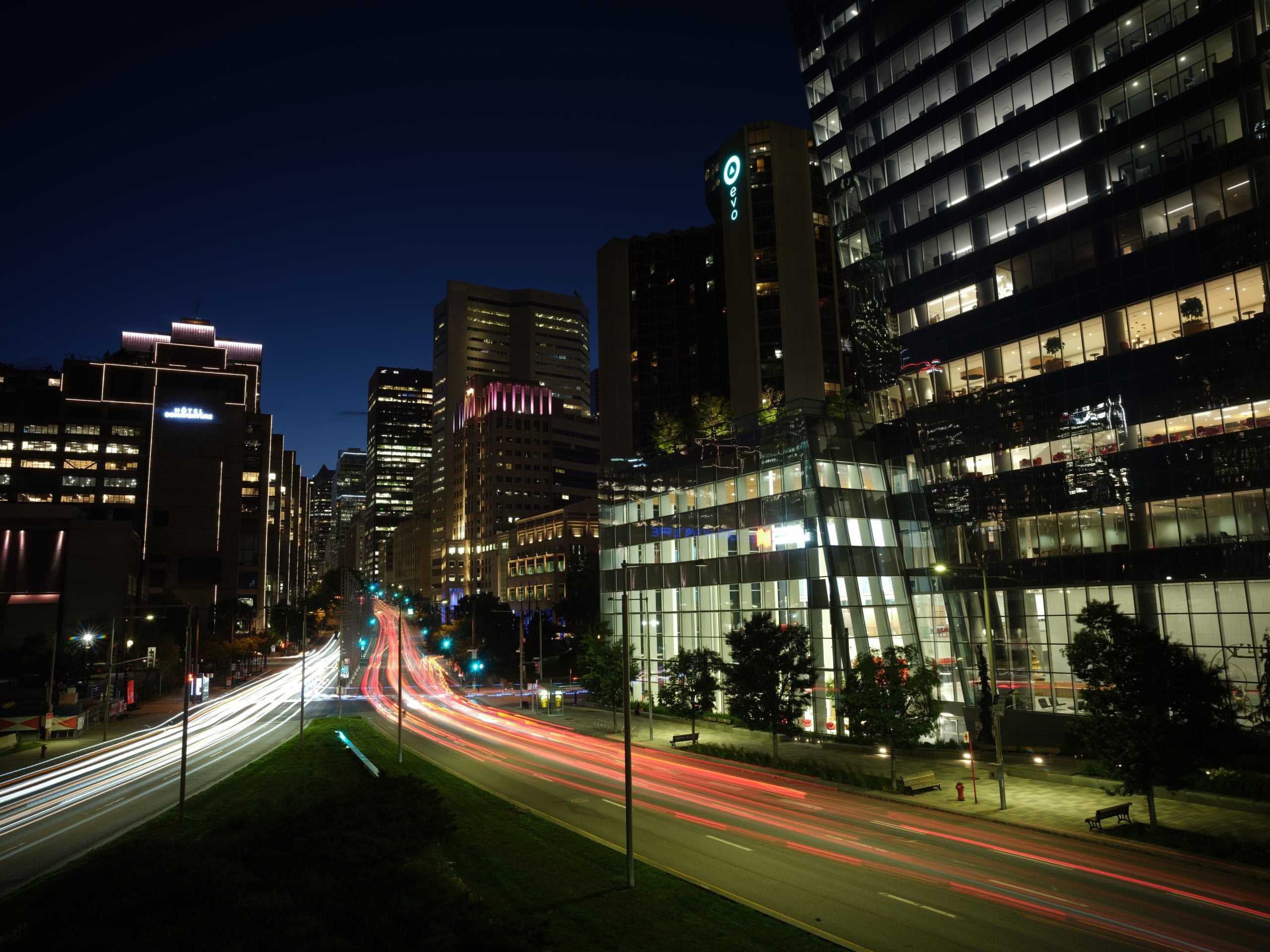 A long-exposure night view of a city street with light trails from moving traffic, surrounded by tall office buildings and hotels.