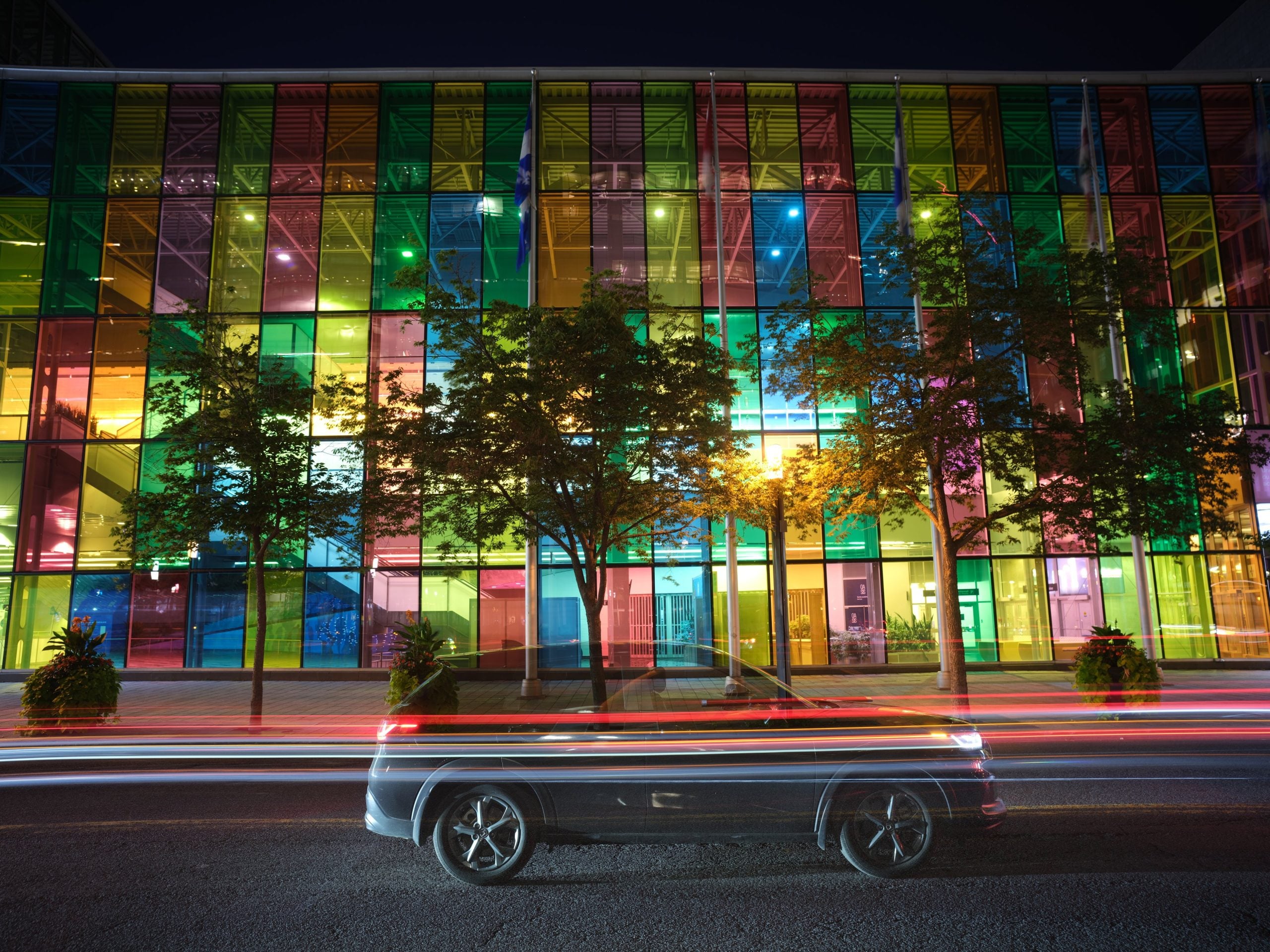 A dark SUV drives past a building with a multi-colored glass facade at night, leaving long light trails from its headlights and taillights.