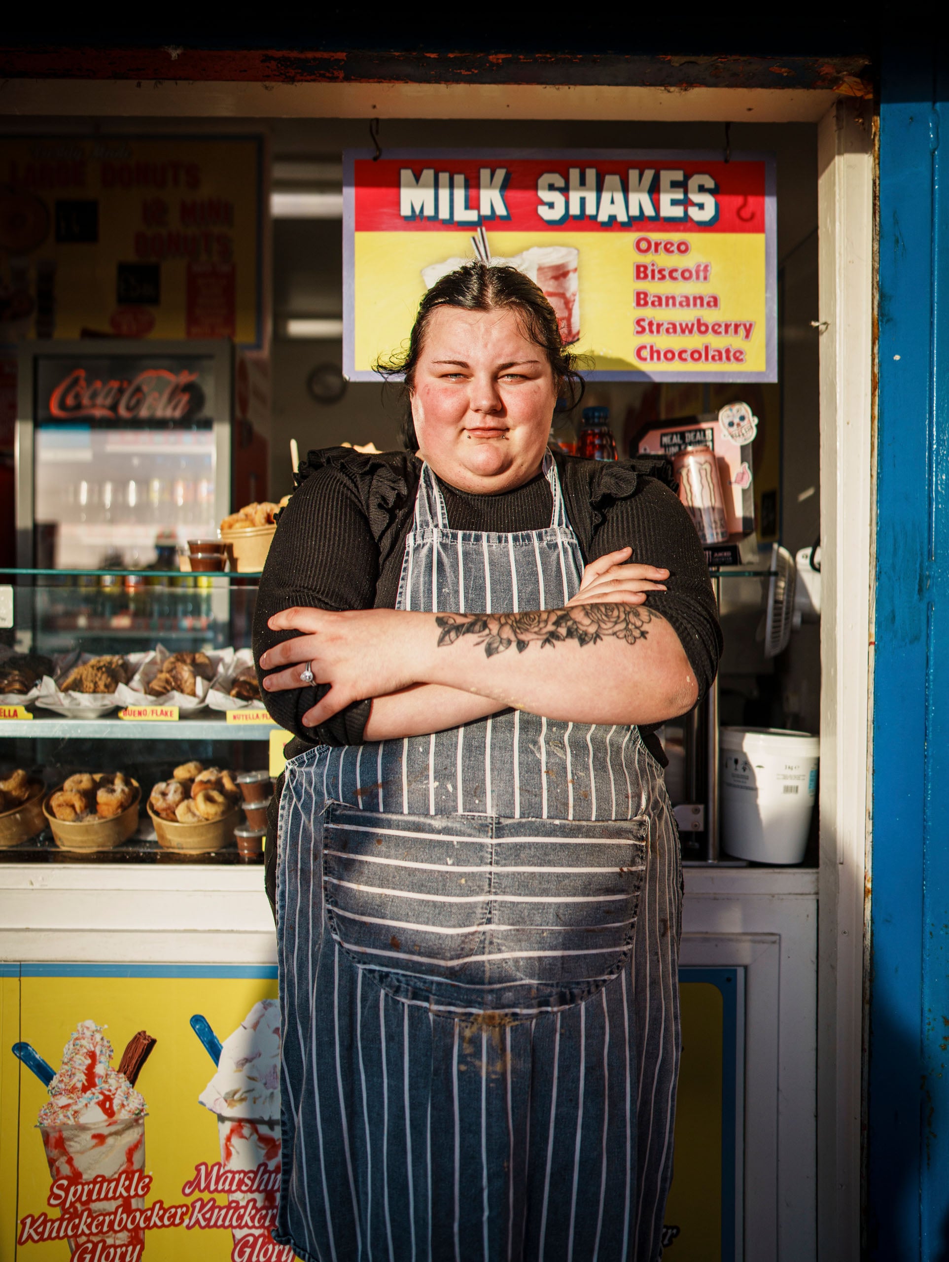 A person with a floral forearm tattoo and a striped apron stands with arms crossed in front of a food stall advertising milkshakes and donuts.