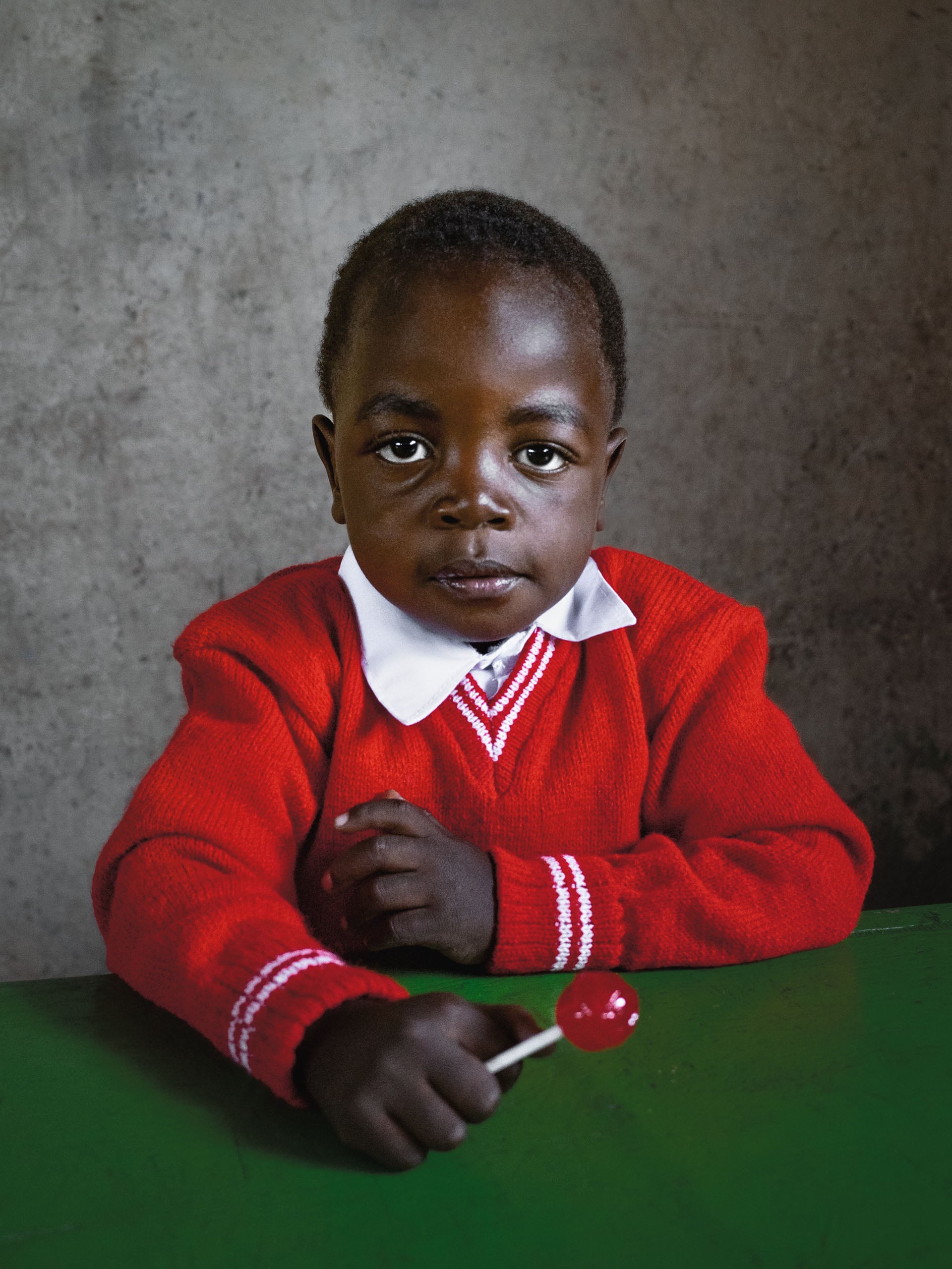 A young child with dark skin wears a red sweater with a white collar and holds a red lollipop while sitting at a green desk.