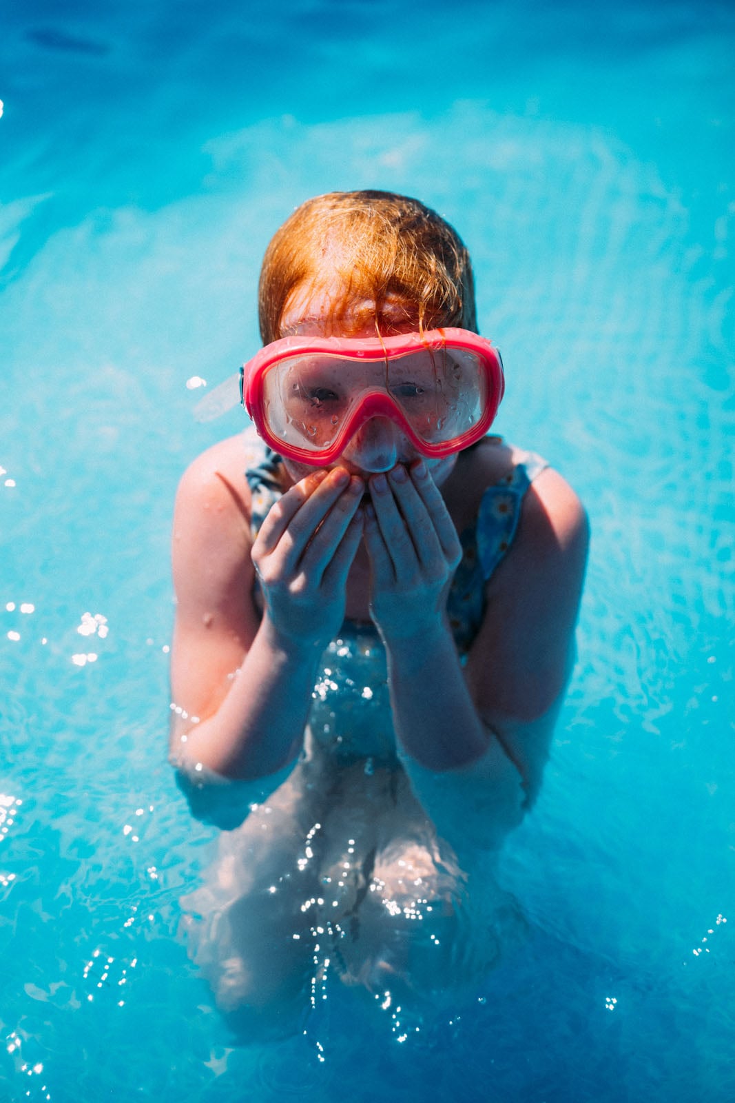 A person with red hair wearing pink swimming goggles stands in a bright blue pool with their hands held up to their face.