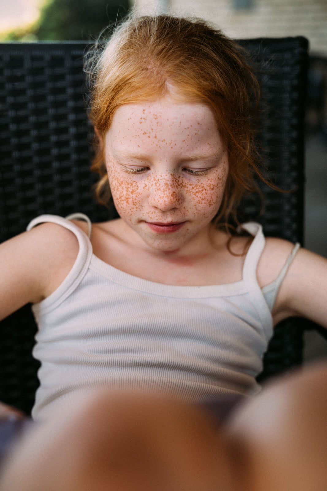 A young girl with red hair and freckles sits with her eyes closed, wearing a white ribbed tank top.