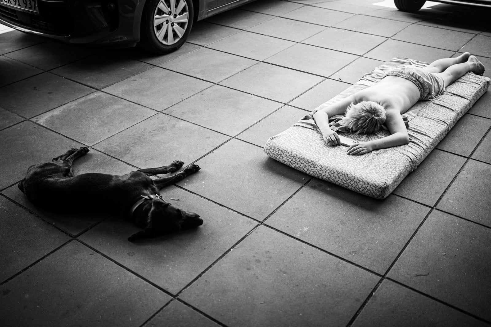 A child lies face down on a thin mattress on a tiled patio next to a black dog resting on the ground.