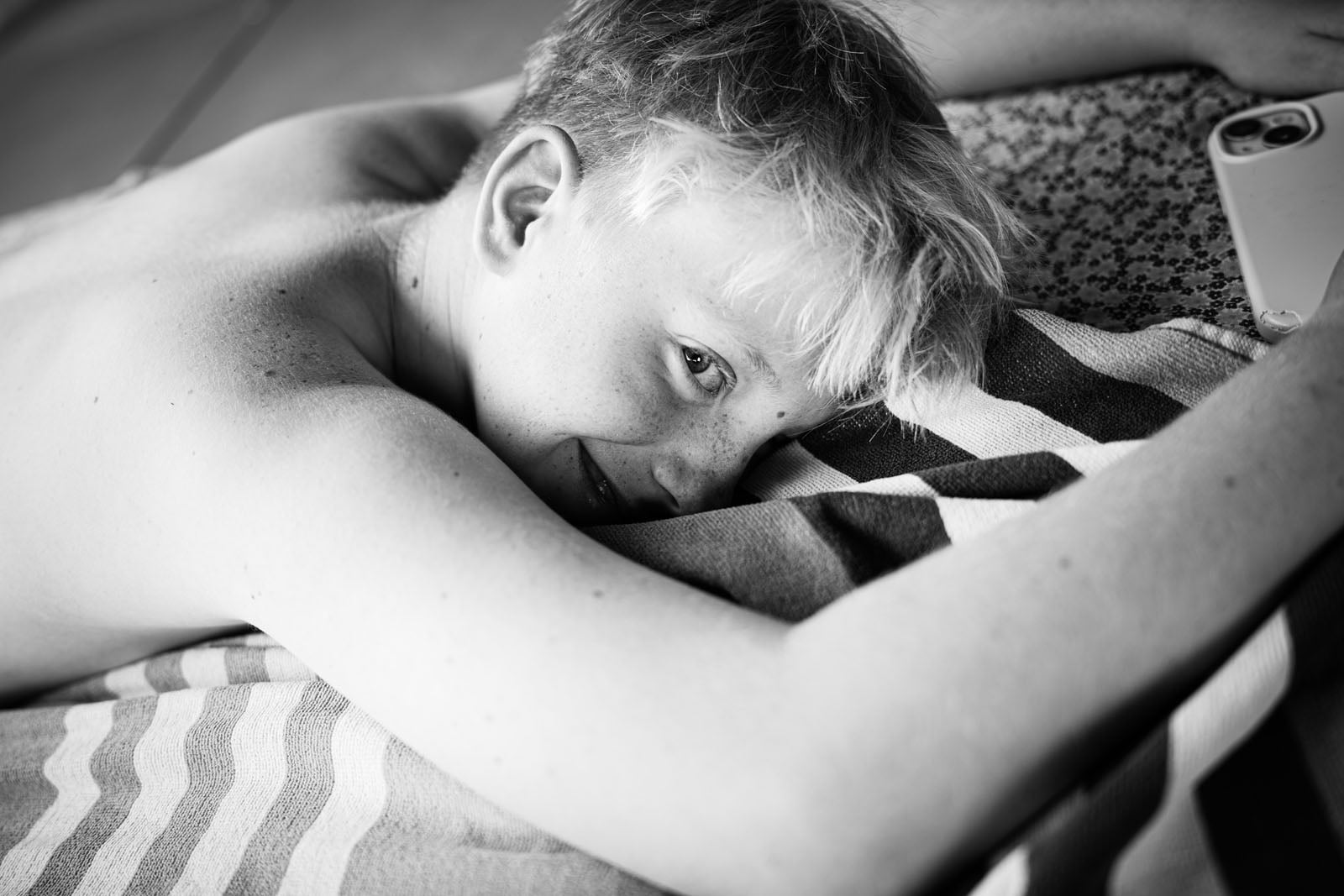 A young person with light hair and freckles lies on their stomach on a striped towel, looking toward the camera while holding a smartphone.