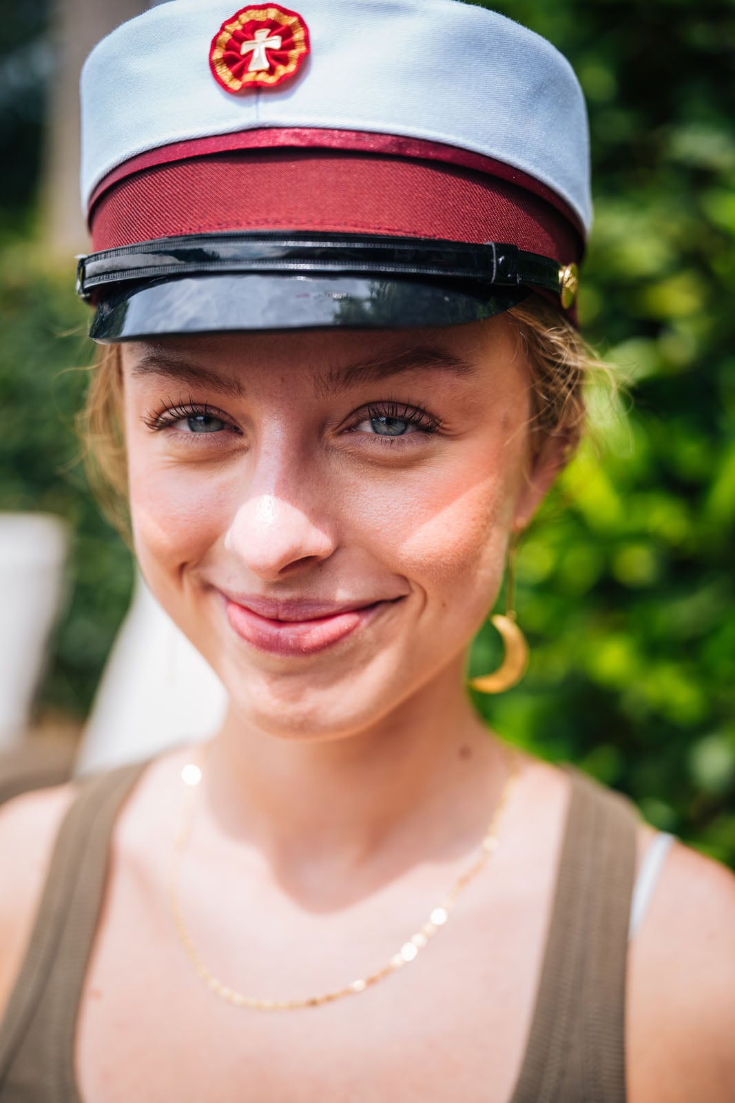 A young woman with light skin and blue eyes smiles at the camera while wearing a traditional Danish student cap and a gold crescent moon earring.