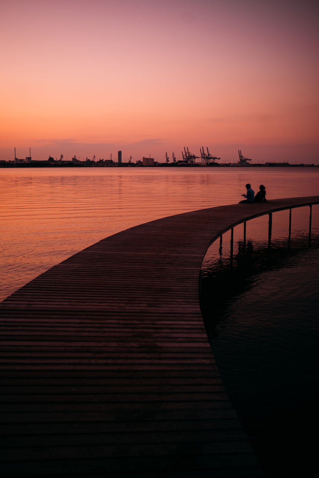Two people sit on a curved wooden pier extending into calm water at sunset, with an industrial port skyline in the distance.