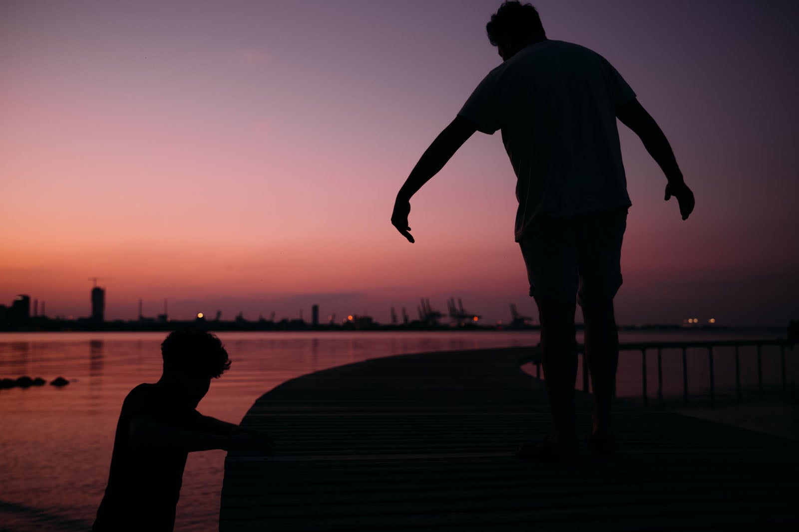 A silhouette of a person standing on a wooden pier reaches toward another person who is partially submerged in the water at sunset.