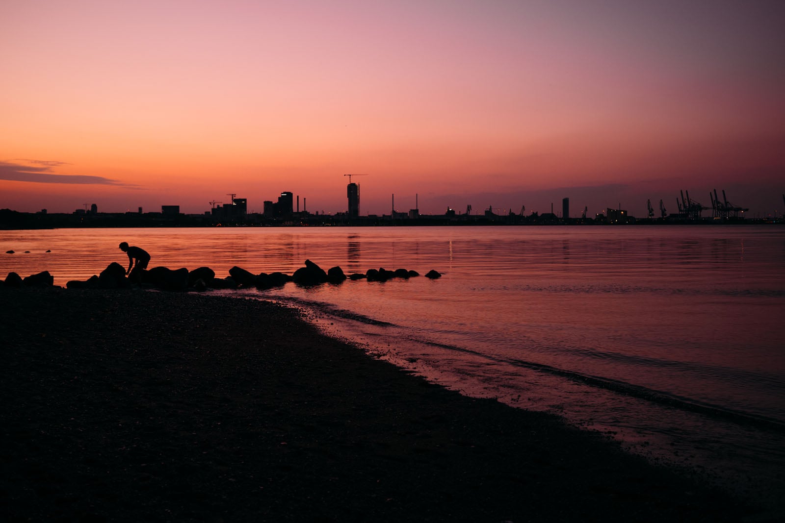 A person stands on a rocky shoreline at sunset, silhouetted against a calm body of water and a distant city skyline.