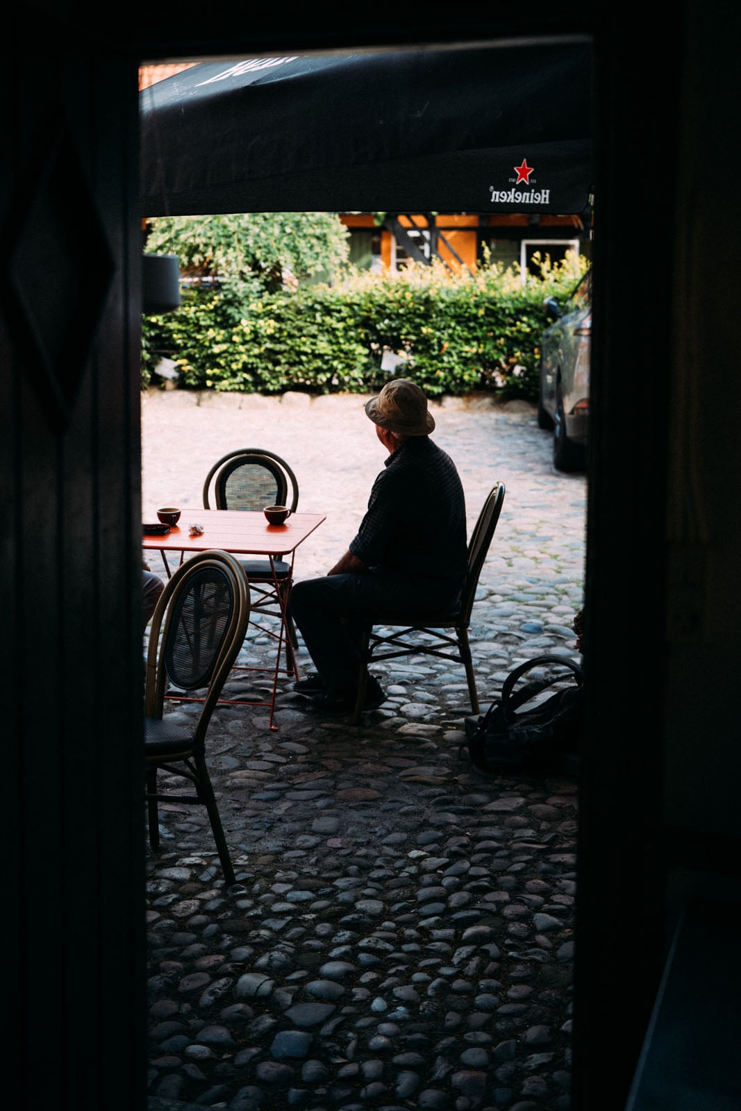 A man in a hat sits at an outdoor table on a cobblestone patio, viewed through a dark doorway.