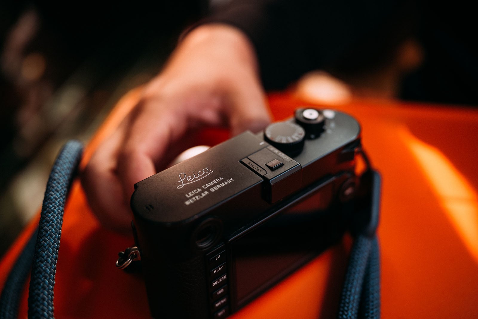 A black Leica camera sits on a bright orange surface, with a hand resting near it and a blue rope strap visible in the foreground.