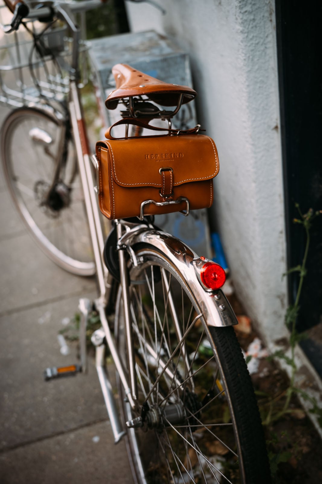 A close-up, high-angle shot of a bicycle parked on a sidewalk, featuring a brown leather saddlebag with the text "HERSKIND" embossed on it.