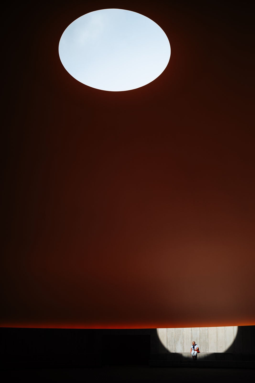 A person sits on a bench within a circular spotlight against a dark, curved wall, beneath a large circular opening in the ceiling.