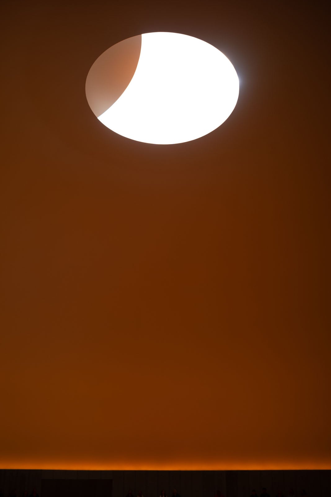 A bright, circular skylight illuminates a large, smooth, orange-toned interior wall, with a dark, indistinct row of people visible at the very bottom.