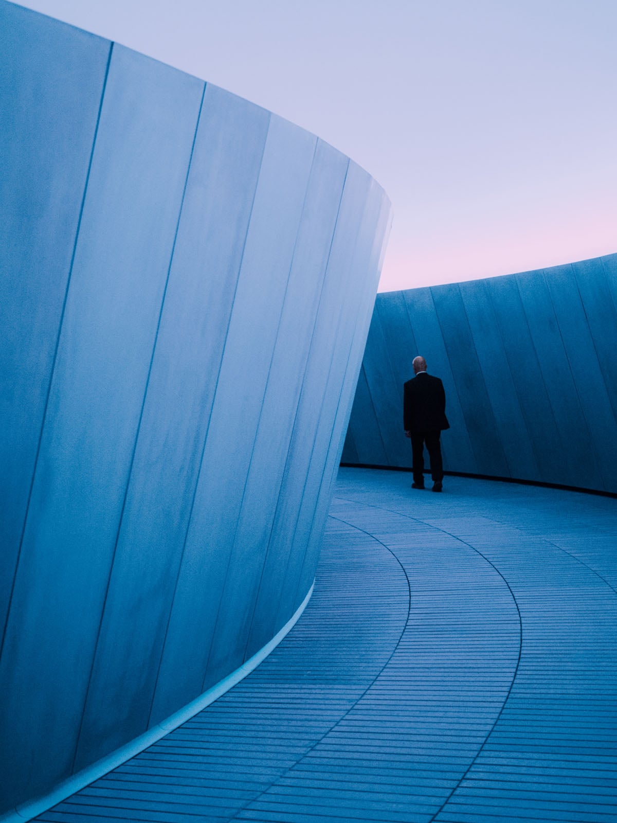 A man in a dark suit walks away along a curved, blue-toned walkway flanked by tall, smooth, panelled walls.