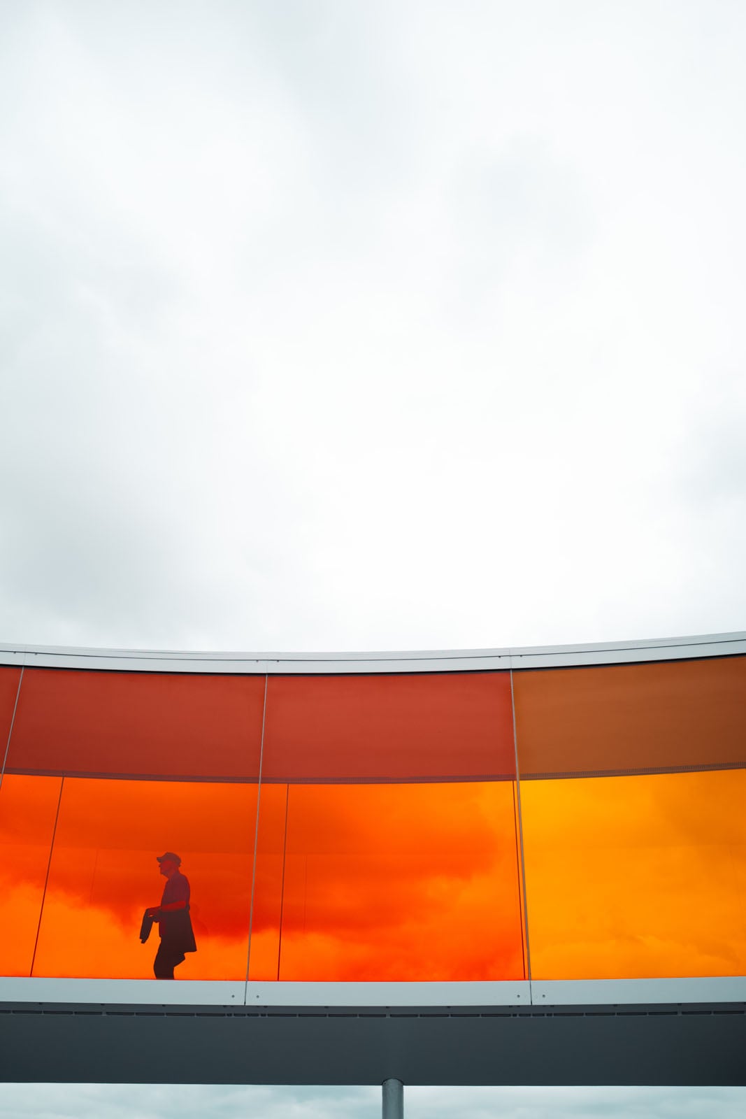 A silhouette of a person walking inside a curved, orange-tinted glass walkway against a cloudy sky.