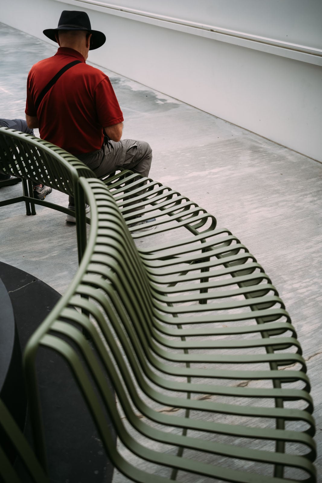 A man wearing a red shirt and a black hat sits on a curved, green metal bench on a concrete surface.