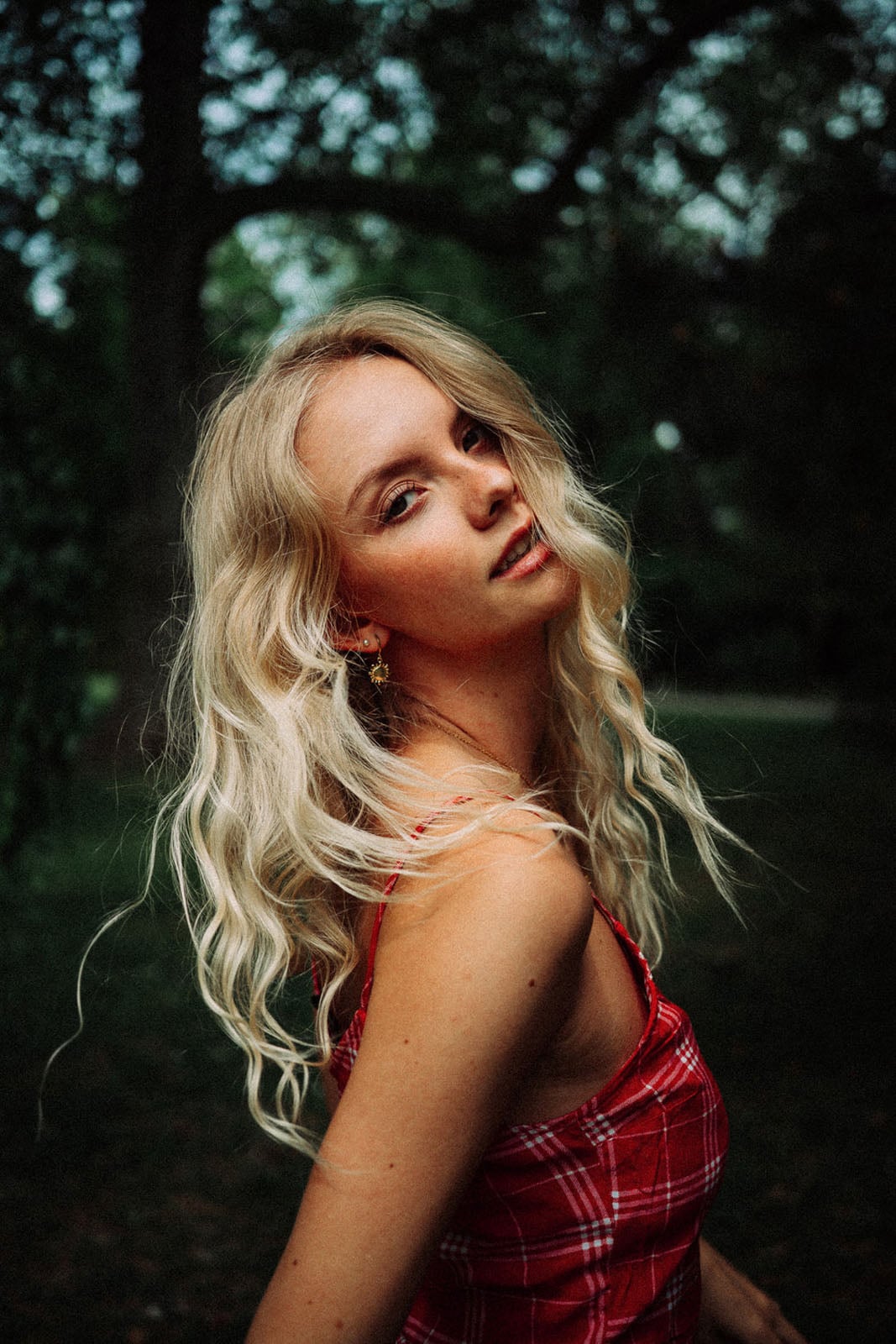A young woman with long, wavy blonde hair and freckles looks over her shoulder while wearing a red plaid top in a wooded area.