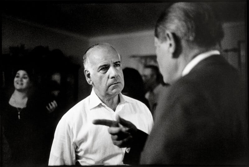 A man in a white shirt looks intently at another man in a dark suit who is pointing a finger toward him, while a woman smiles in the background.