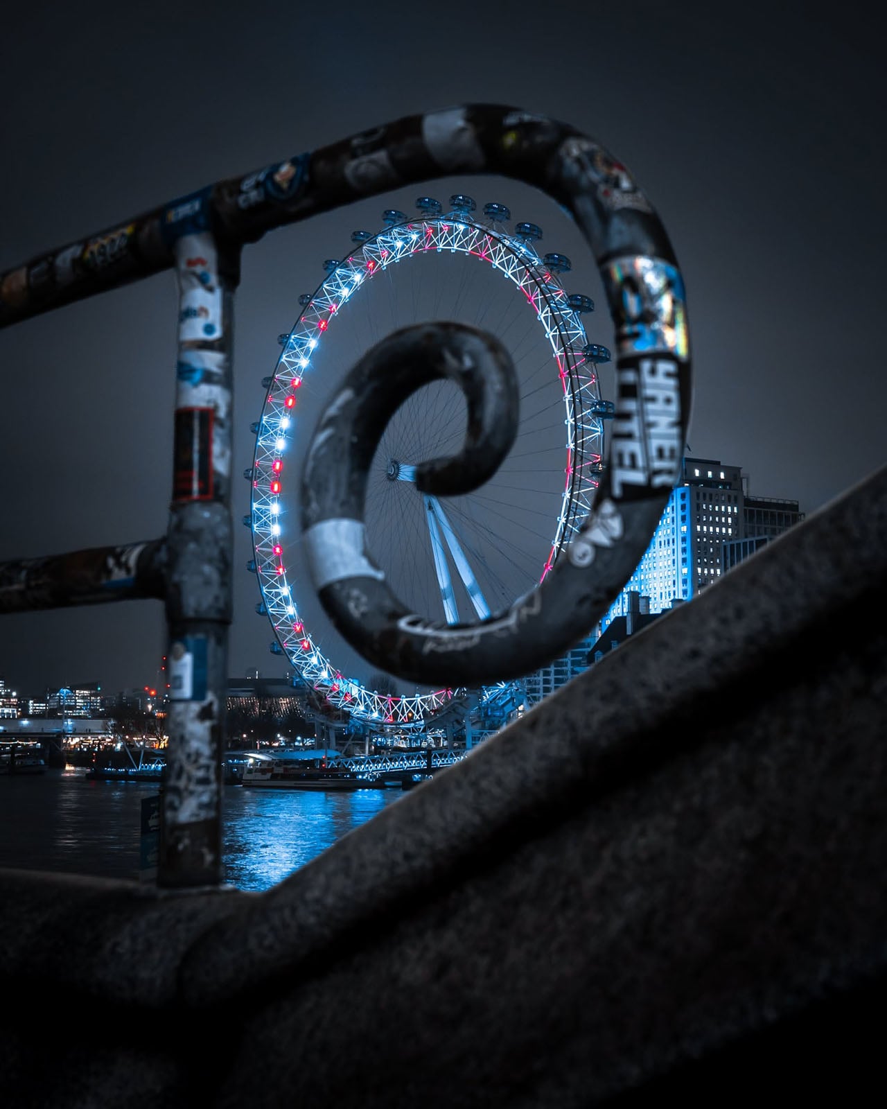 The London Eye illuminated at night, viewed through the spiral metal railing of a bridge covered in stickers.