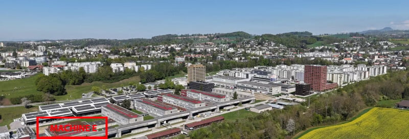 An aerial view of a sprawling industrial complex with flat-roofed buildings, surrounded by a residential town and green hills under a clear blue sky, with a red-outlined label reading "MACHINE E" in the foreground.