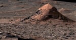 A large, conical rock formation sits in a vast, rocky Martian landscape covered in scattered stones and wind-swept sand dunes.